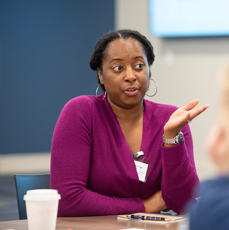 A woman in a purple sweater speaks during a small group discussion at a professional event, gesturing with one hand while seated at a table with a coffee cup and notebook.