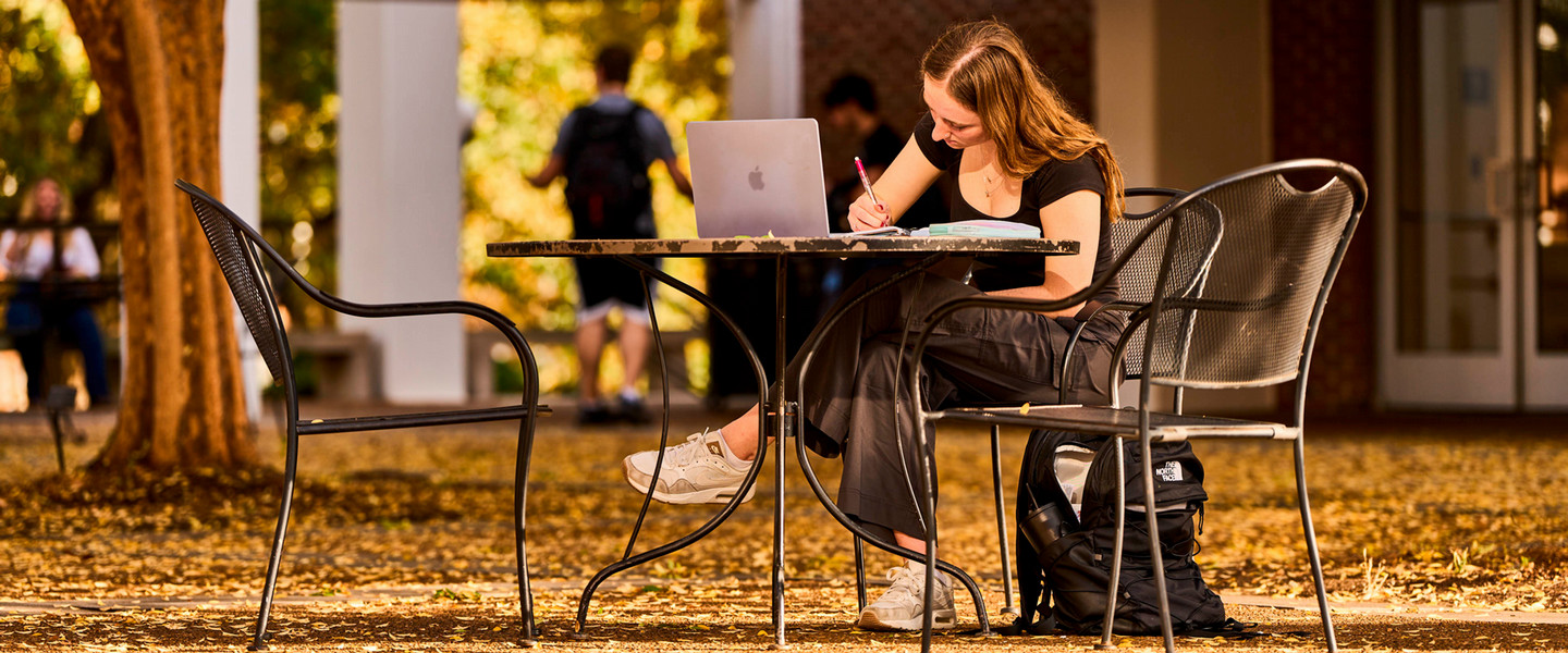 A student sitting a a table outside writing in a notebook with her laptop in front of her