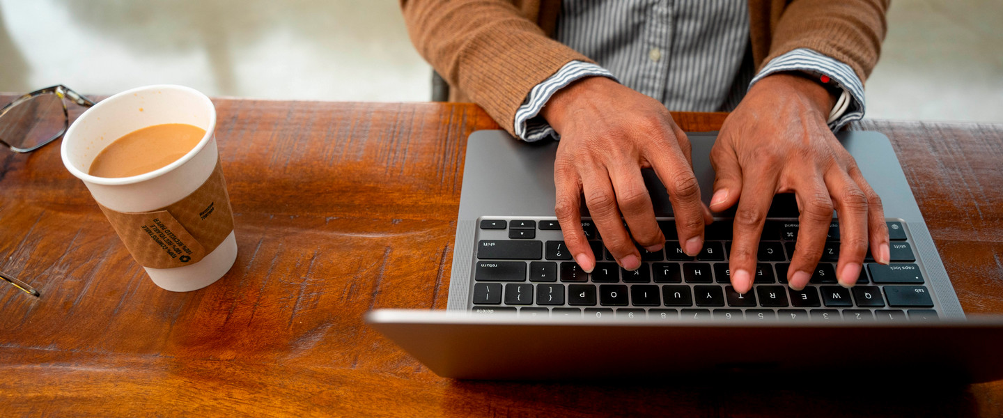 Above shot of woman's hands typing on a laptop with a cup of coffee to the left