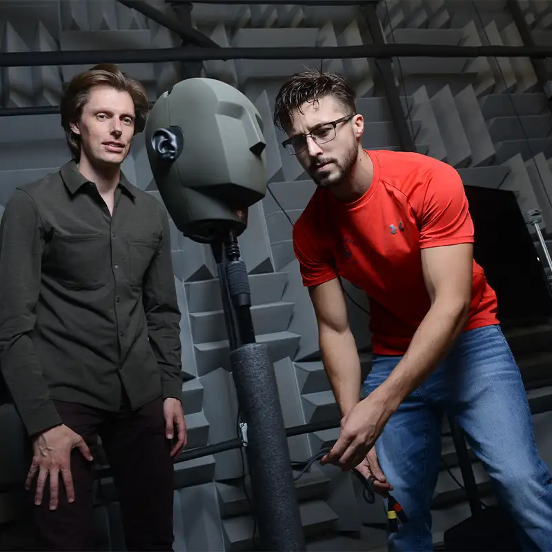 An indoor photo of two men standing around a testing dummy inside of a soundproof room. 