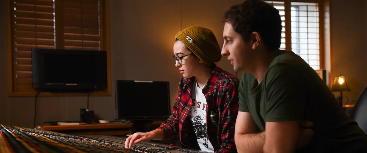 A woman wearing glasses and a beanie, dressed in a plaid shirt, is adjusting a mixing console alongside a man with short hair and a green shirt. They are both focused on the controls in a music studio with soft lighting.