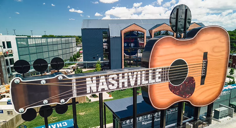 Large outdoor guitar-shaped sign with the word “Nashville” across the fretboard, overlooking modern buildings beneath a bright blue sky.