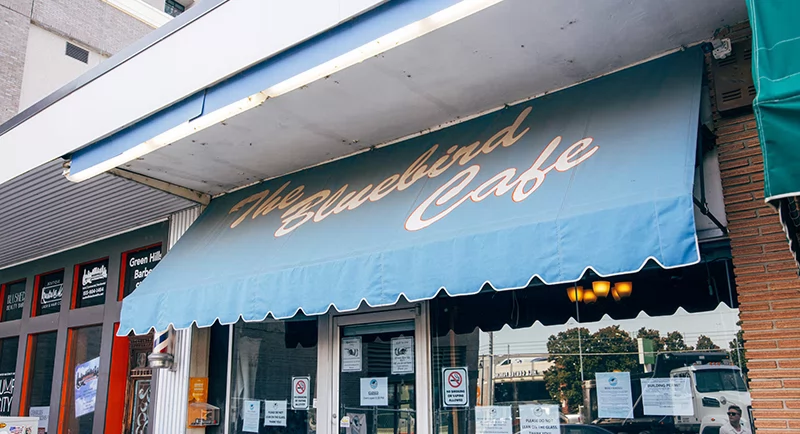 Front entrance of The Bluebird Café with its blue awning, glass door, posted signs, and neighboring storefronts.