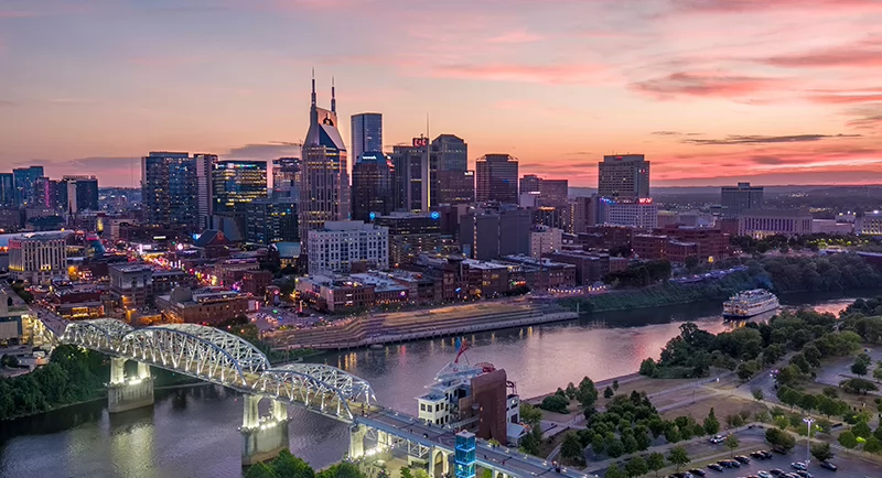 Aerial view of downtown Nashville at sunset with colorful sky, river reflections, bridges, and illuminated city buildings.