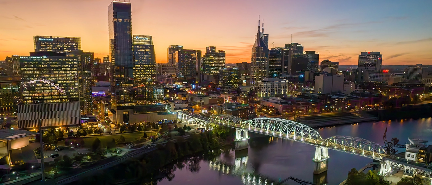erial view of downtown Nashville at sunset, with city skyscrapers lit up and the John Seigenthaler Pedestrian Bridge reflecting across the Cumberland River.
