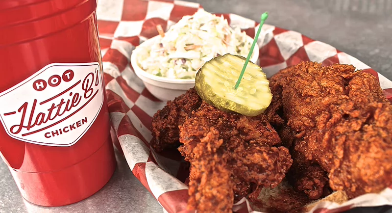Basket of spicy fried chicken topped with pickle slices, served with coleslaw and a red Hattie B’s cup on a checkered liner.