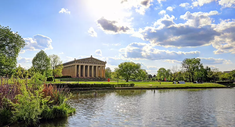 Wide view of the Parthenon in Centennial Park on a sunny day, with bright clouds, green trees, and a calm pond in the foreground.