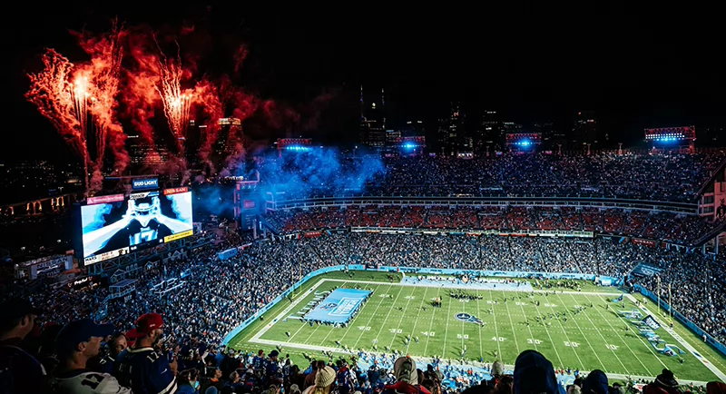 Nighttime football game at Nissan Stadium with a full crowd, bright lights, and fireworks bursting over the field.
