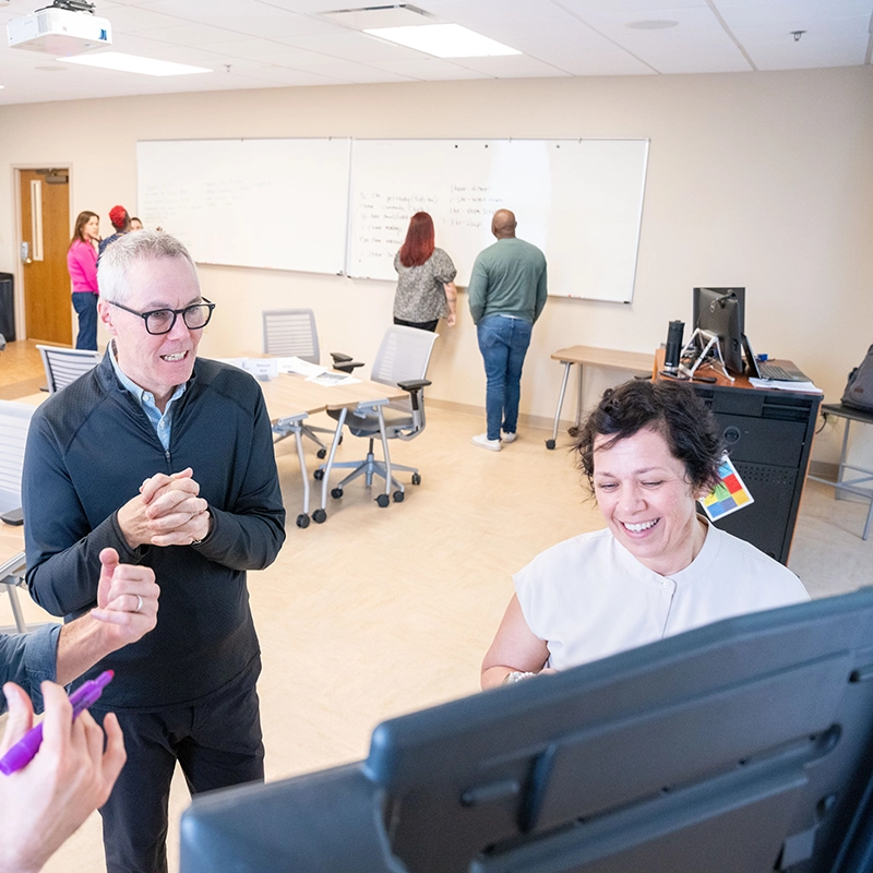 A man with gray hair, glasses, and a black pullover clasps his hands while talking to a smiling woman with short dark hair in a white shirt seated in front of a monitor. Someone in the foreground holds a purple marker, and in the background, other group members stand writing on large whiteboards during a collaborative training session.