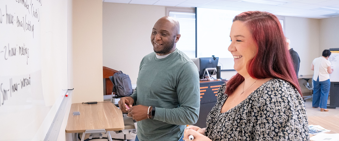 A smiling woman with long reddish-brown hair and a floral top stands next to a man wearing a long-sleeved green sweater. Both are holding dry-erase markers while looking at handwritten notes on a large whiteboard in a bright seminar room or office setting.