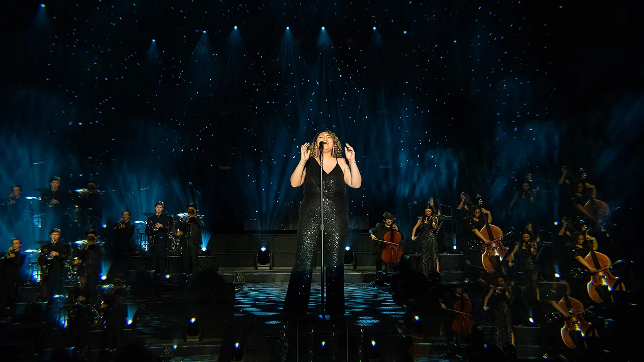 A powerful vocal performance by a female singer with a live orchestra on stage at the Best of the Best Showcase, set against a backdrop of starry lighting effects.