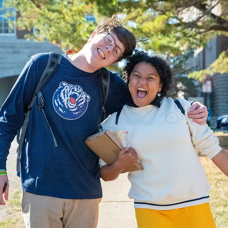 Two students smiling and posing outdoors, one with a backpack and the other holding books, enjoying a sunny day on campus.