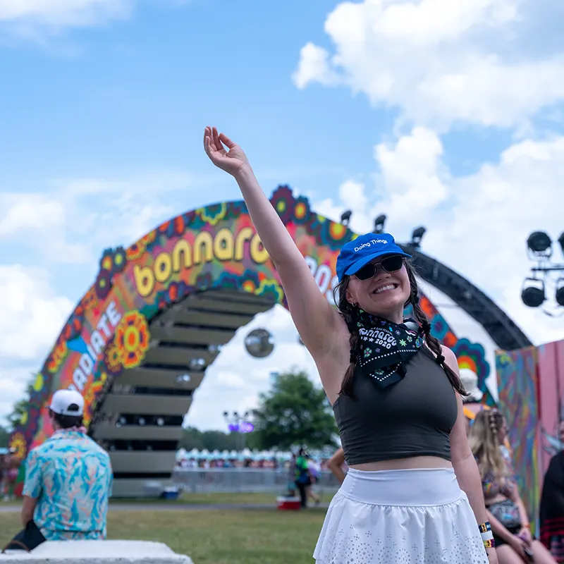 A smiling person raises their hand in front of the colorful Bonnaroo festival entrance, enjoying the outdoor event.