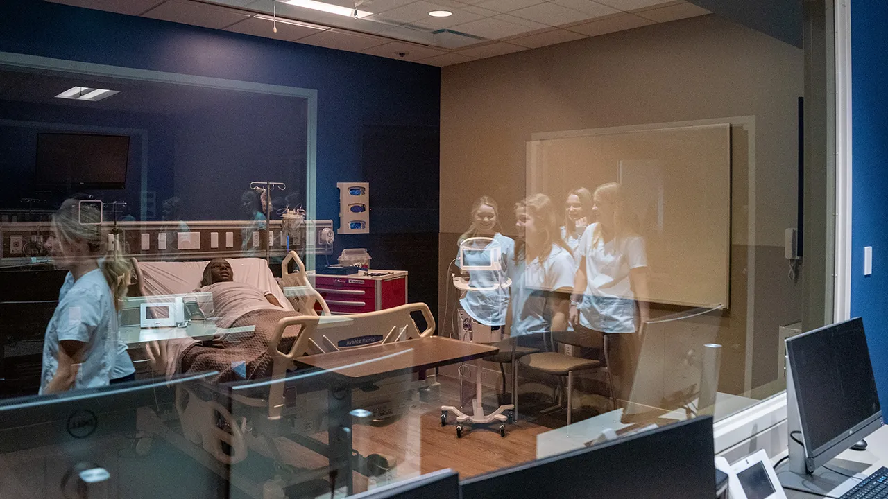 Students observing a patient in a hospital bed through a window in a simulation lab, with medical equipment and monitors in the foreground.