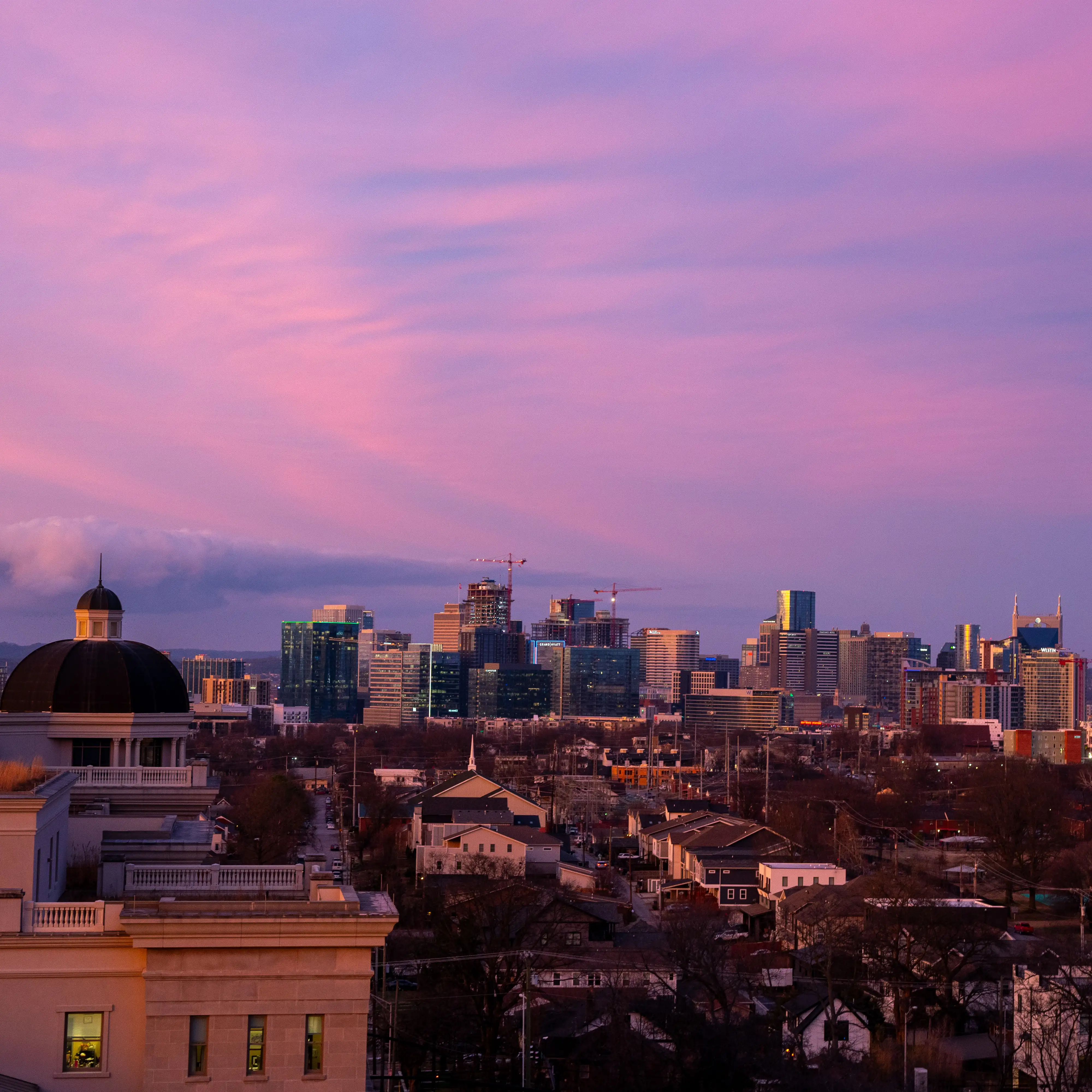 A wide view of the Nashville skyline at sunset, with the sky glowing pink and purple above a mix of modern high-rise buildings and older neighborhood homes in the foreground. The domed roof of a Belmont University campus building appears on the left.