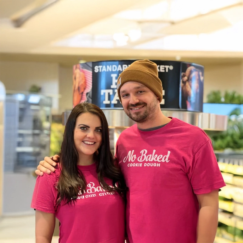 Megan and Jimmy Feeman, founders of No Baked Cookie Dough, smiling and standing together in a store, both wearing matching pink No Baked t-shirts.