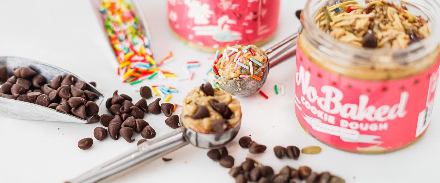 A close-up image of No Baked Cookie Dough jars surrounded by scoops of cookie dough topped with chocolate chips and colorful sprinkles, alongside scattered chocolate chips and a jar of rainbow sprinkles.