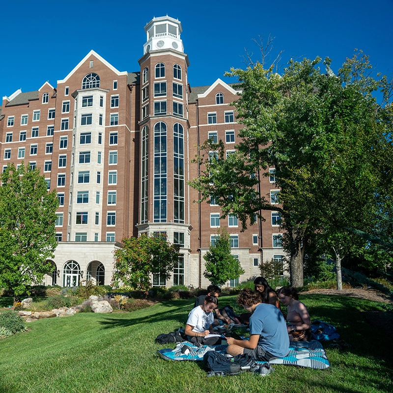 A group of students sits on a grassy lawn outside a tall, brick residence hall on Belmont's campus with large windows and a prominent tower, enjoying the sunny day while studying and socializing.