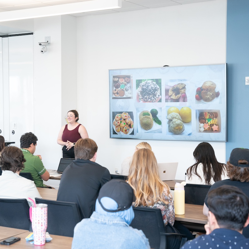 A woman giving a presentation to a group of students in a classroom, with a screen displaying images of baked goods, including cookies, pastries, and cakes.