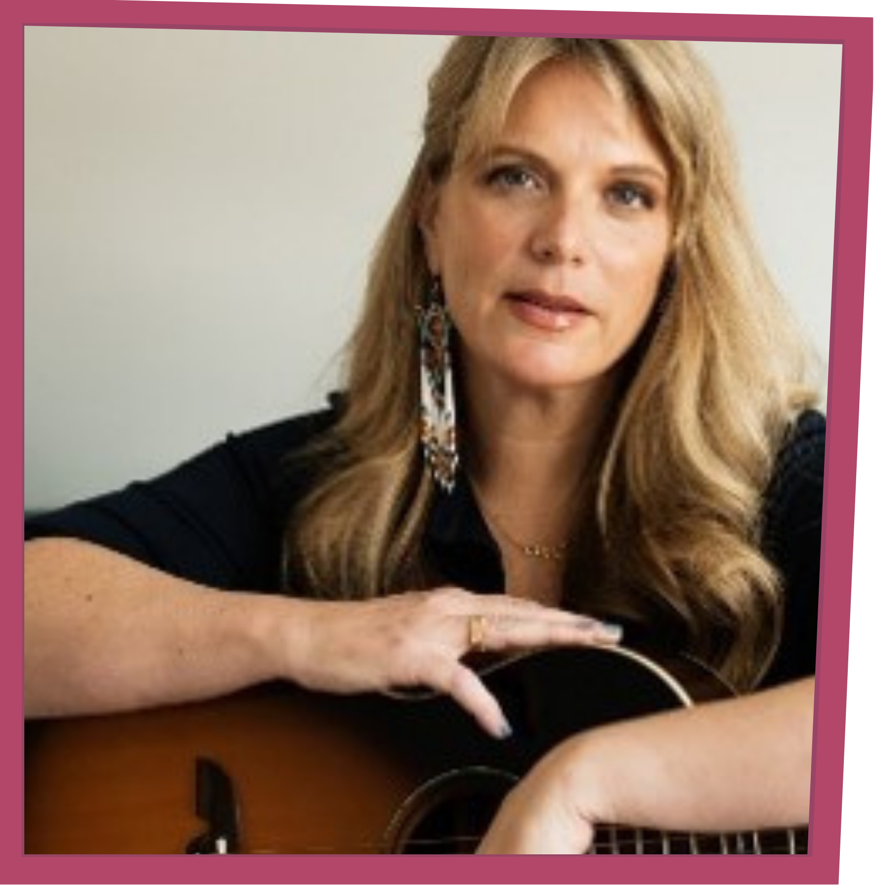 Portrait of a woman with long blonde hair resting her arms on an acoustic guitar, wearing a dark top and a long dangling earring.