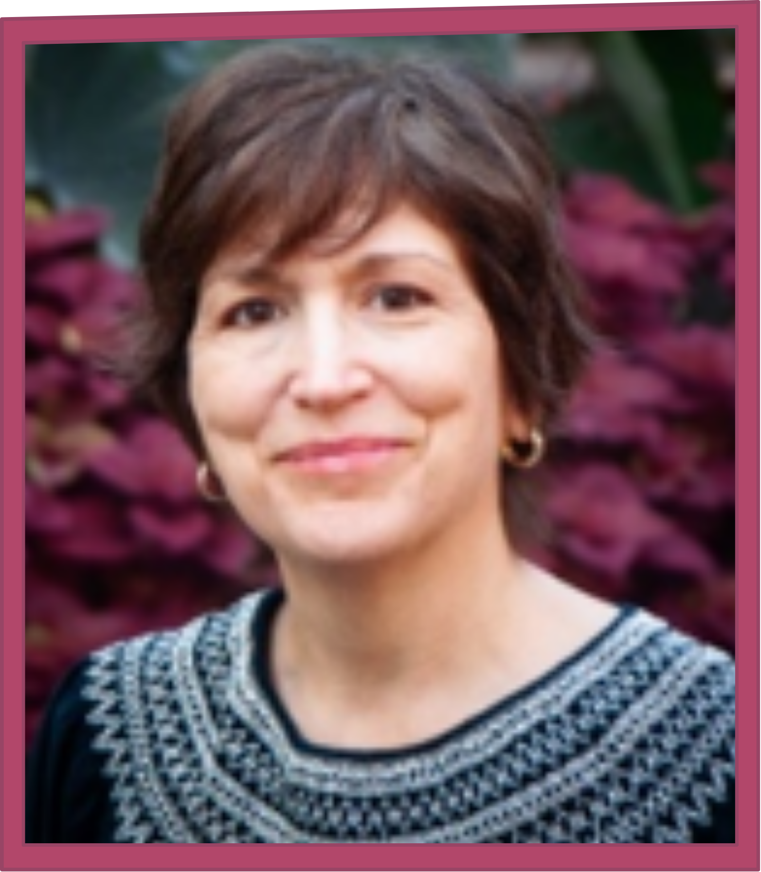 Head-and-shoulders portrait of a smiling person with short brown hair, wearing small hoop earrings and a patterned sweater, with blurred purple foliage in the background.