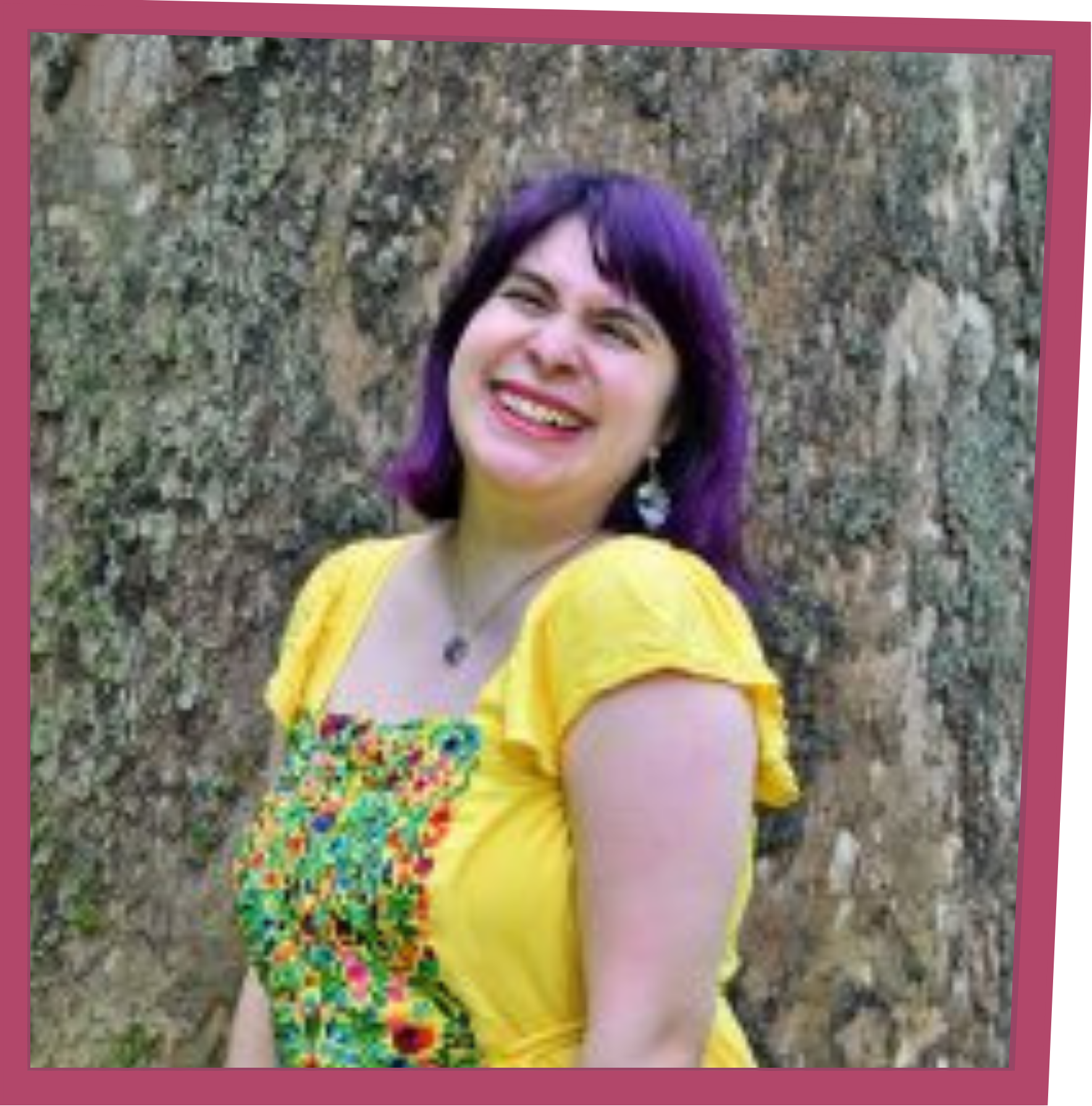 Smiling person with purple hair wearing a yellow top with a colorful floral pattern, standing in front of a rock wall.