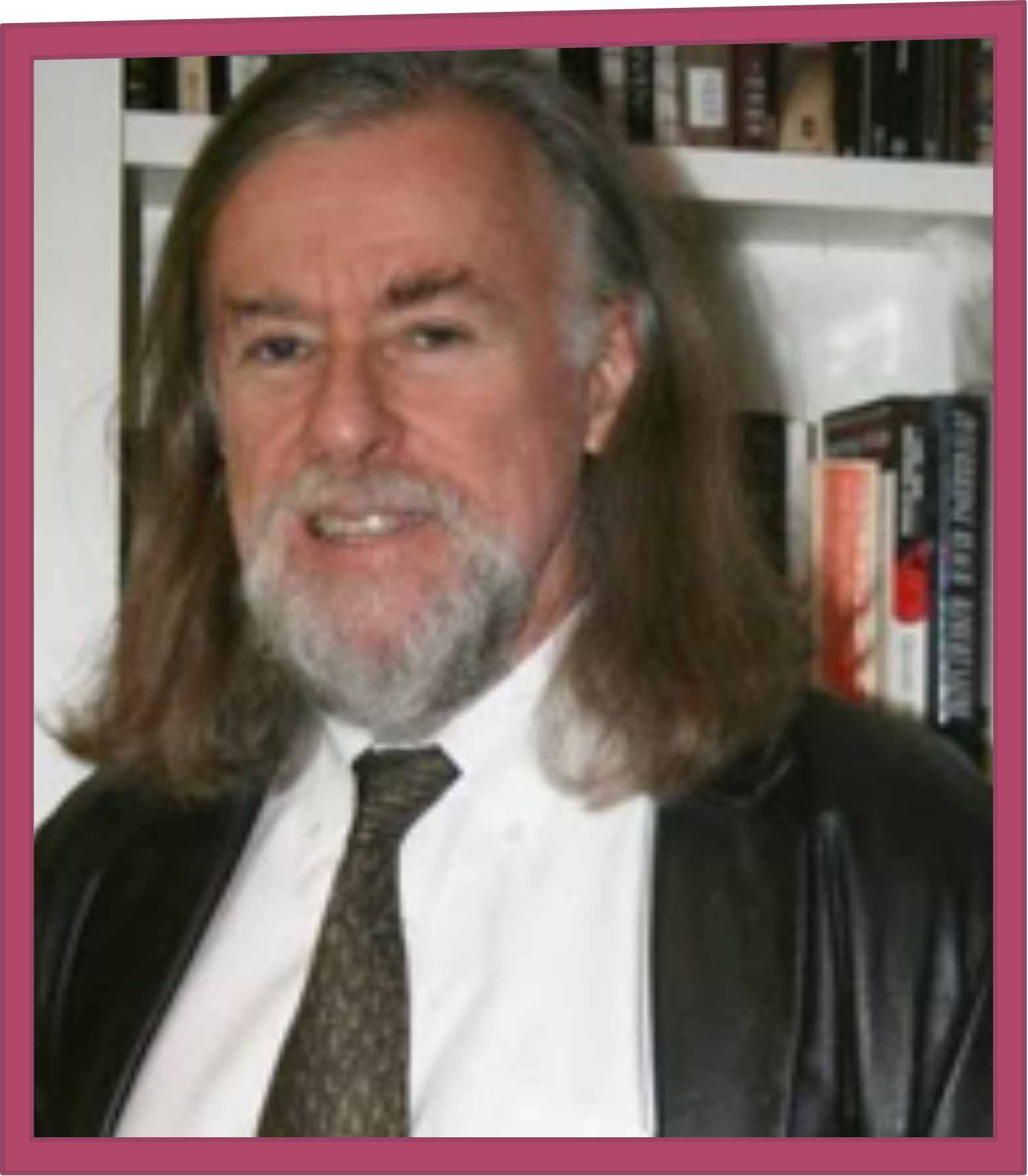 Portrait of a long-haired, bearded man in a suit and tie, standing in front of bookshelves.