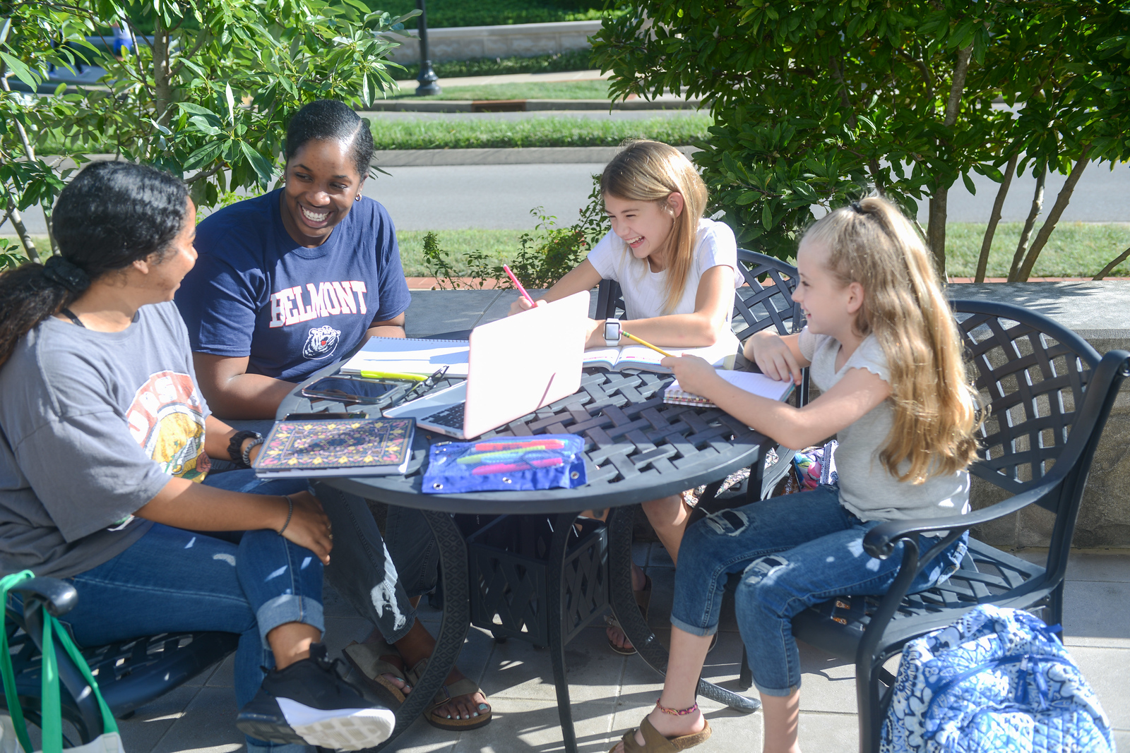Belmont College of Education students and children sitting outdoors at a table, smiling and working together with notebooks and a laptop.