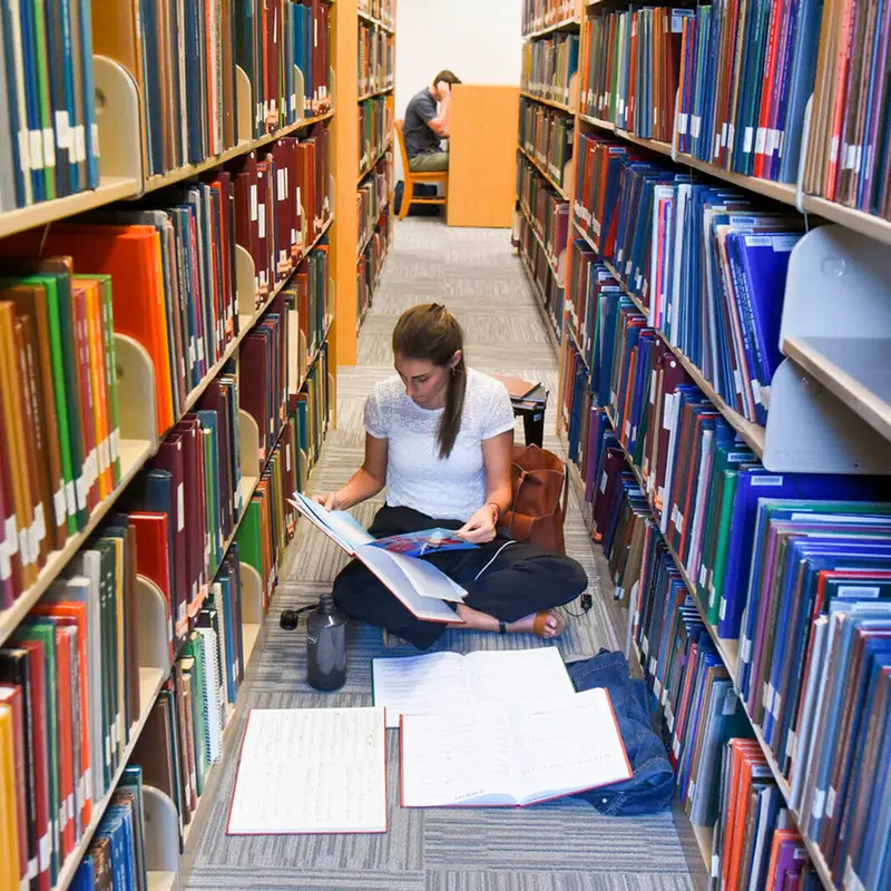 Student sitting on the floor between the bookshelves studying surrounded by books