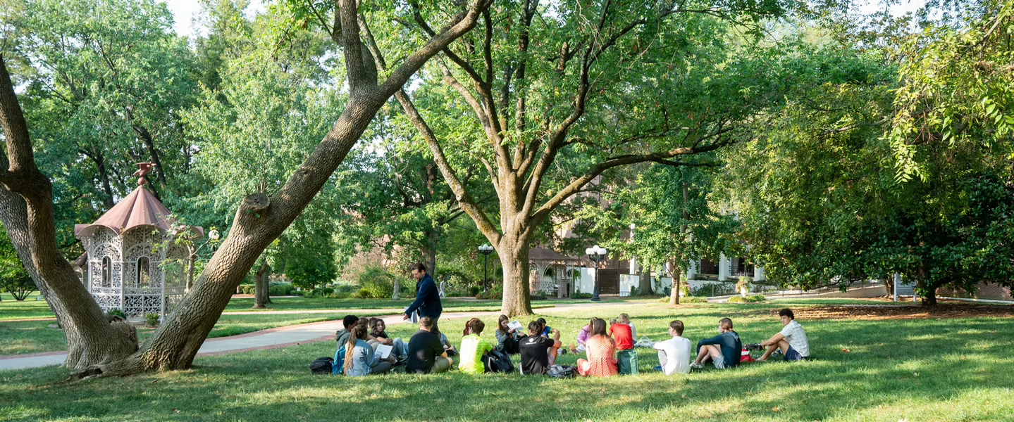 Students sitting on the lawn on the historic side of campus under trees during class outside