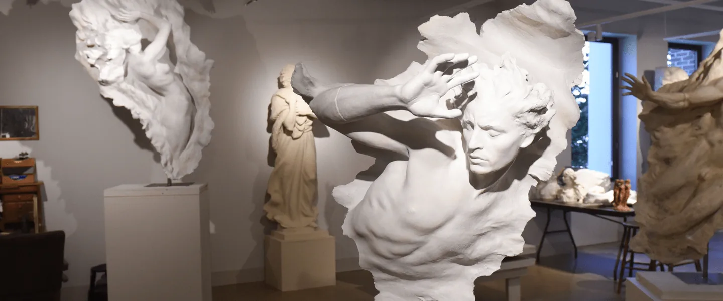 Three large white plaster figures dynamically emerging from rough backdrops on display inside the Frederick Hart Studio Museum, with a studio desk visible in the background.