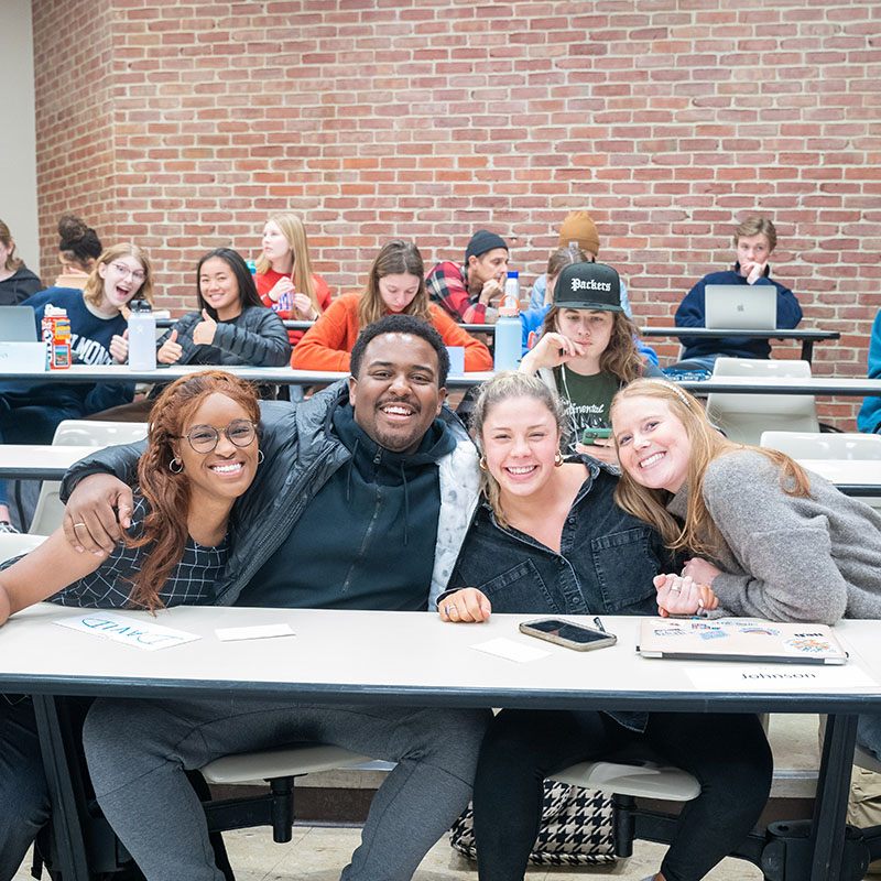 four students in class hugging and smiling at the camera