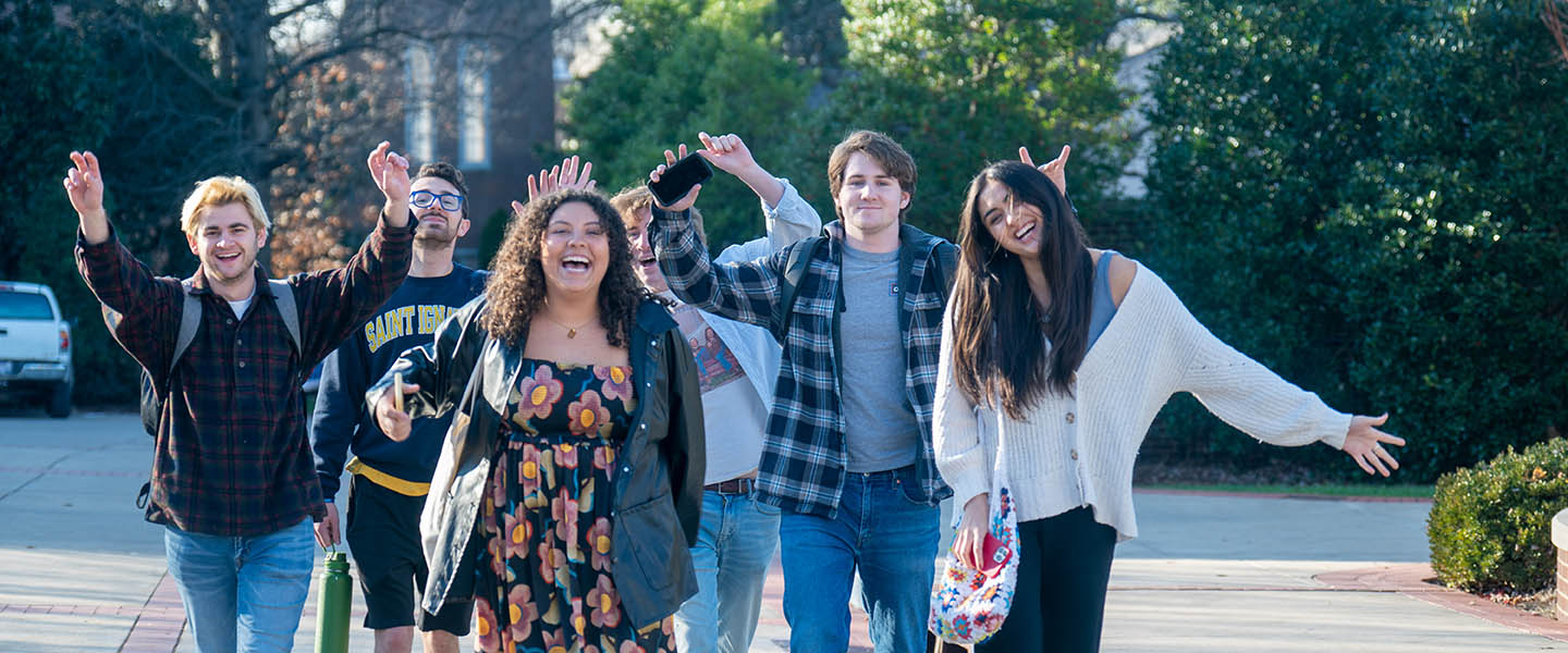 Six students outside walking, smiling, and waving 