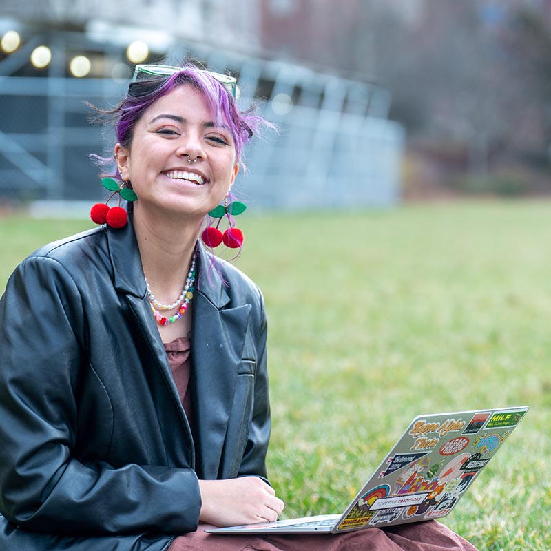 Female student sitting on the grass with a laptop in her lap while she smiles at the camera