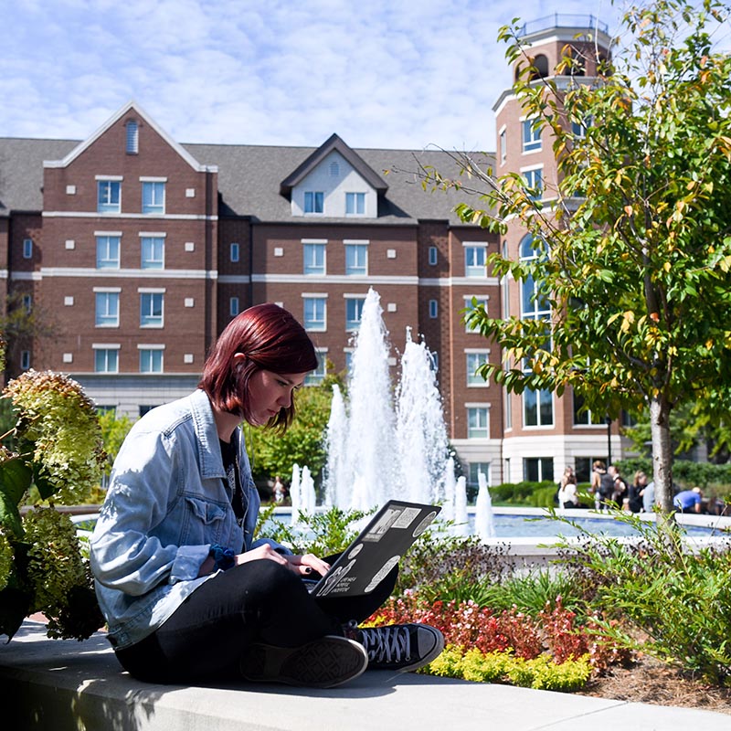 Female student sitting on a ledge in front of a fountain with a laptop in her lap