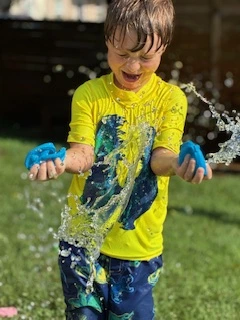 Child in a yellow shirt laughing while playing with water balloons as water splashes in a grassy yard.