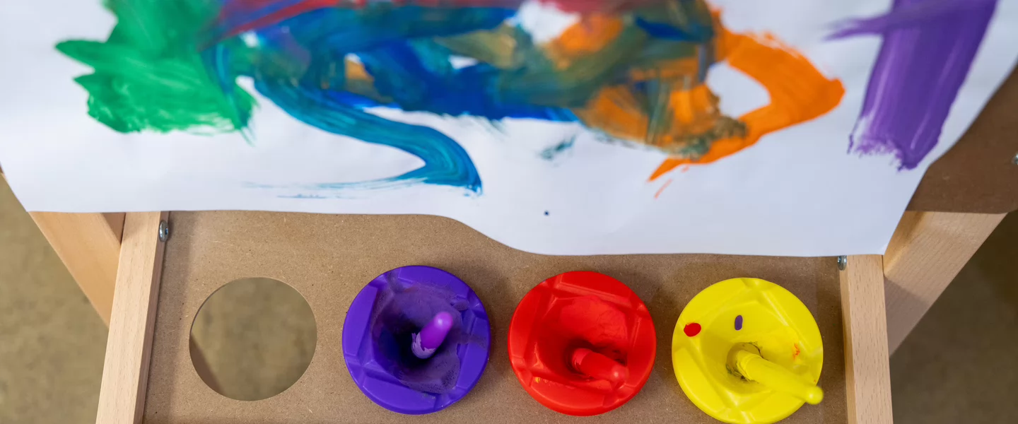 Children’s paint cups and brushes beneath a sheet of colorful finger painting.