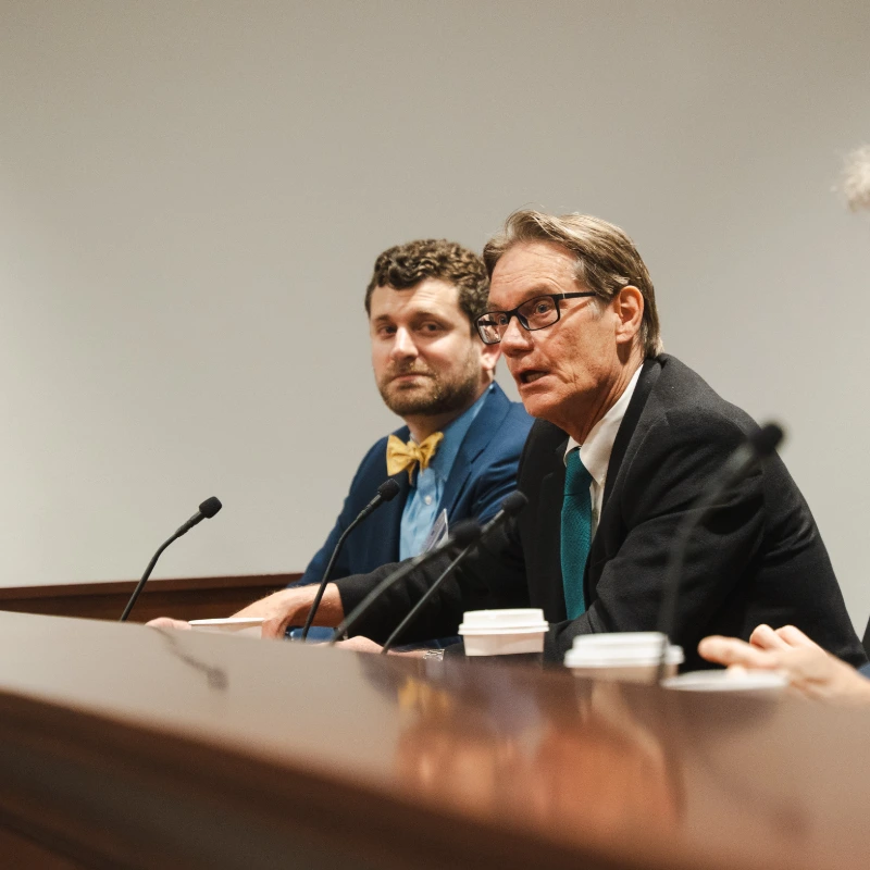 Two men at a panel discussion: an older man in glasses speaking, a younger man with a beard and yellow bow tie.