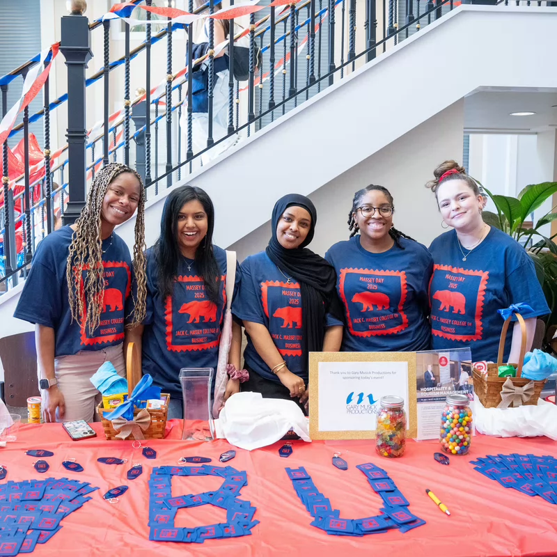 Five students stand behind a vibrant table for Massey Day, smiling warmly. The table is decorated with red and blue items, including a sign that reads "#BU."