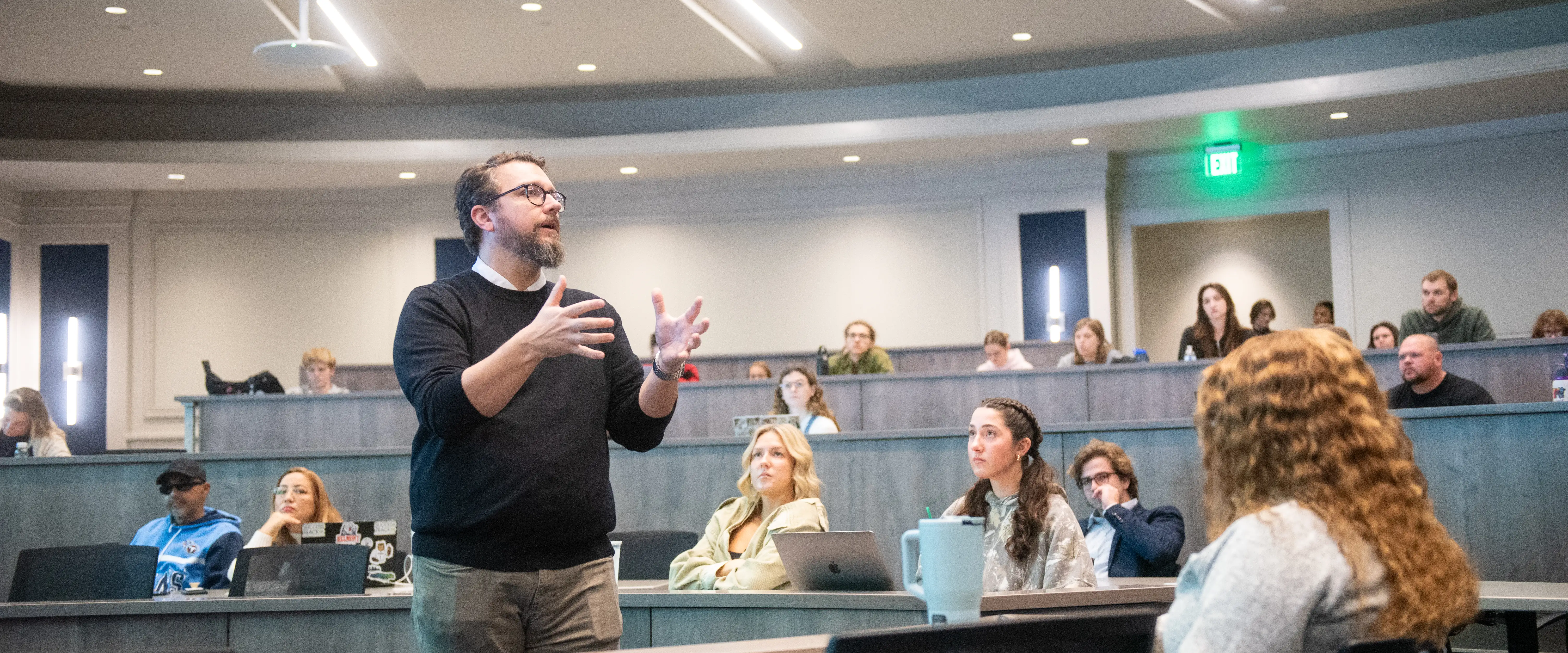 Professor speaking and gesturing during a classroom discussion with students seated in a tiered lecture hall.