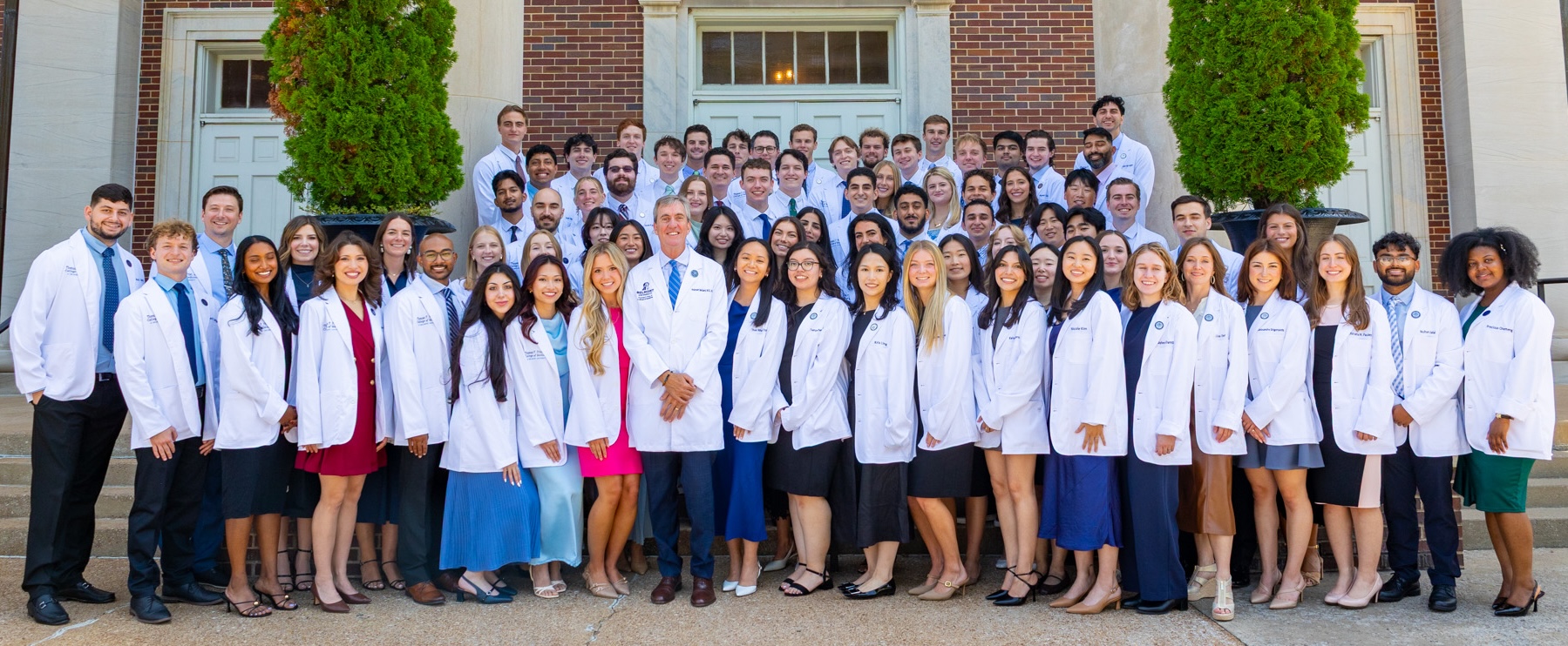The Thomas F. Frist, Jr. College of Medicine at Belmont University Class of 2030 poses for a group photo on the steps of a campus building. The students, wearing white lab coats, stand together smiling and looking towards the camera. 