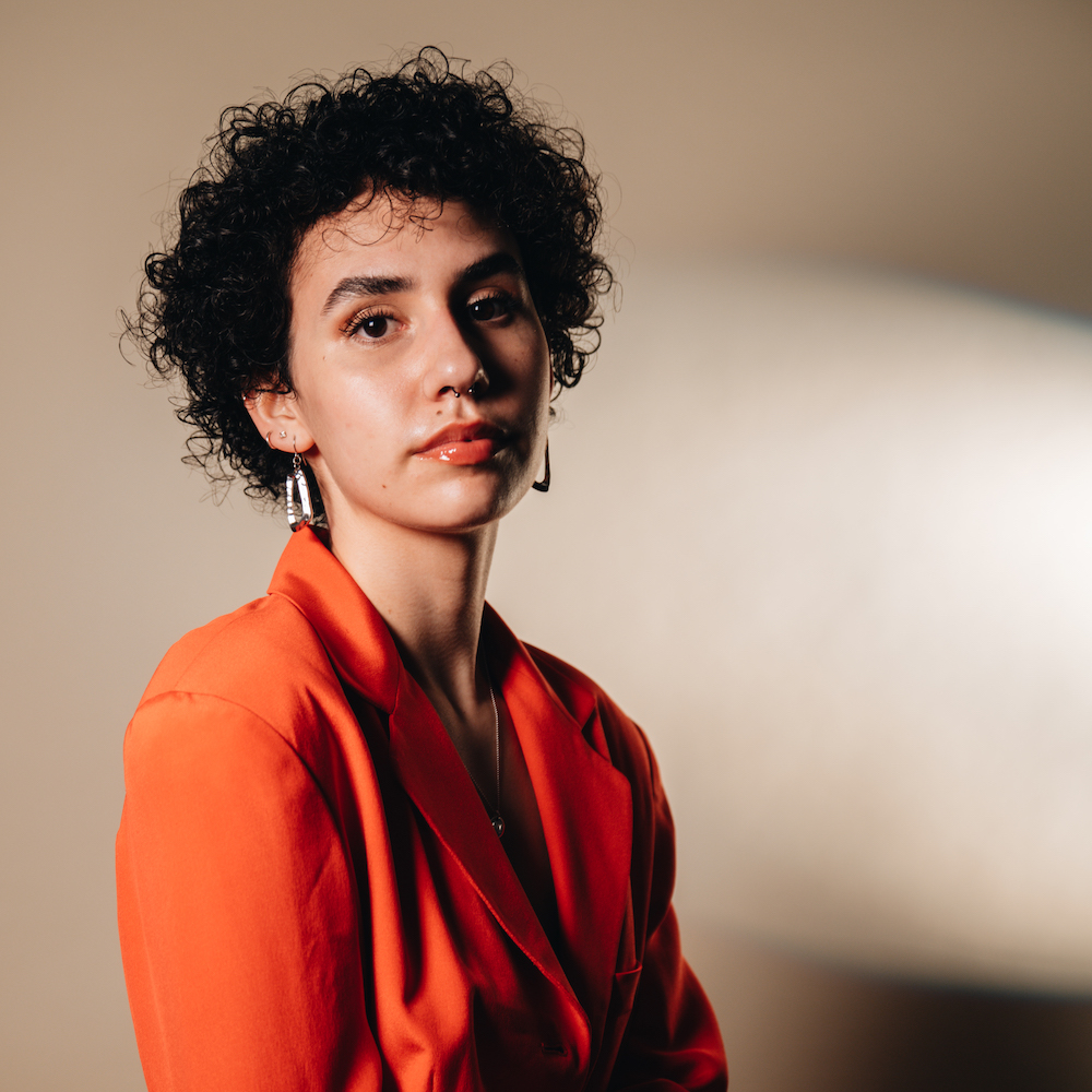 Professional portrait of Marian Anderson, featuring her with short curly hair, wearing a bold orange blazer and silver hoop earrings. She poses confidently against a softly lit neutral background.
