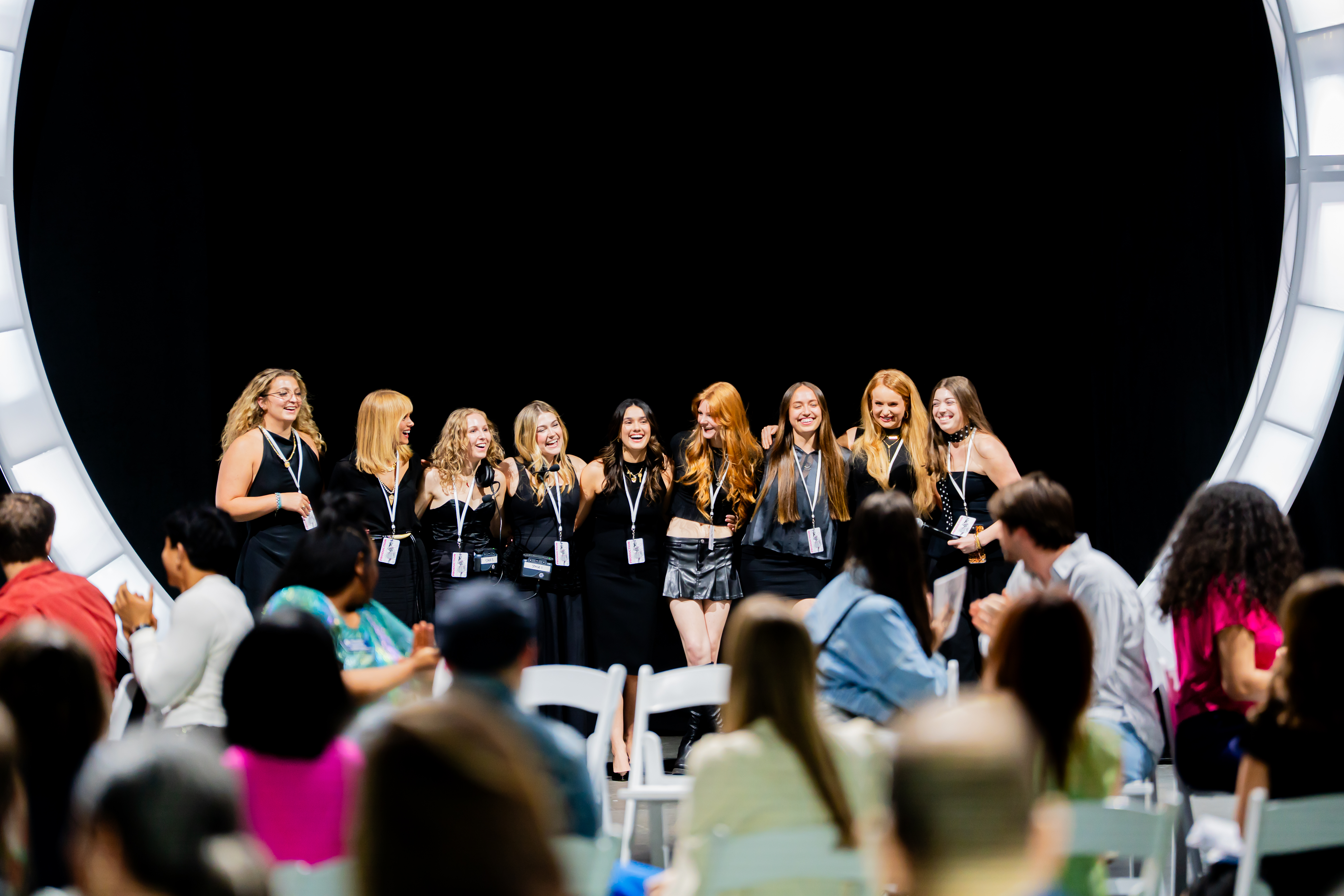 Models walk down a brightly lit runway at a fashion show, with the student designer waving to the audience. The venue is dimly lit except for the stage lights overhead, which highlight the models and their outfits. Two large screens on either side of the runway display a live feed of the show. The audience, seated along both sides of the runway, watches and applauds.