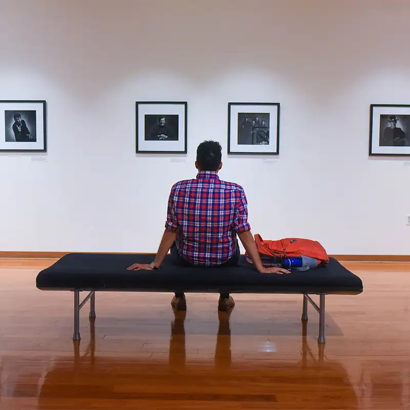 Person seated on a bench in an art gallery, facing a row of framed black-and-white photographs on the wall, with a red backpack beside them