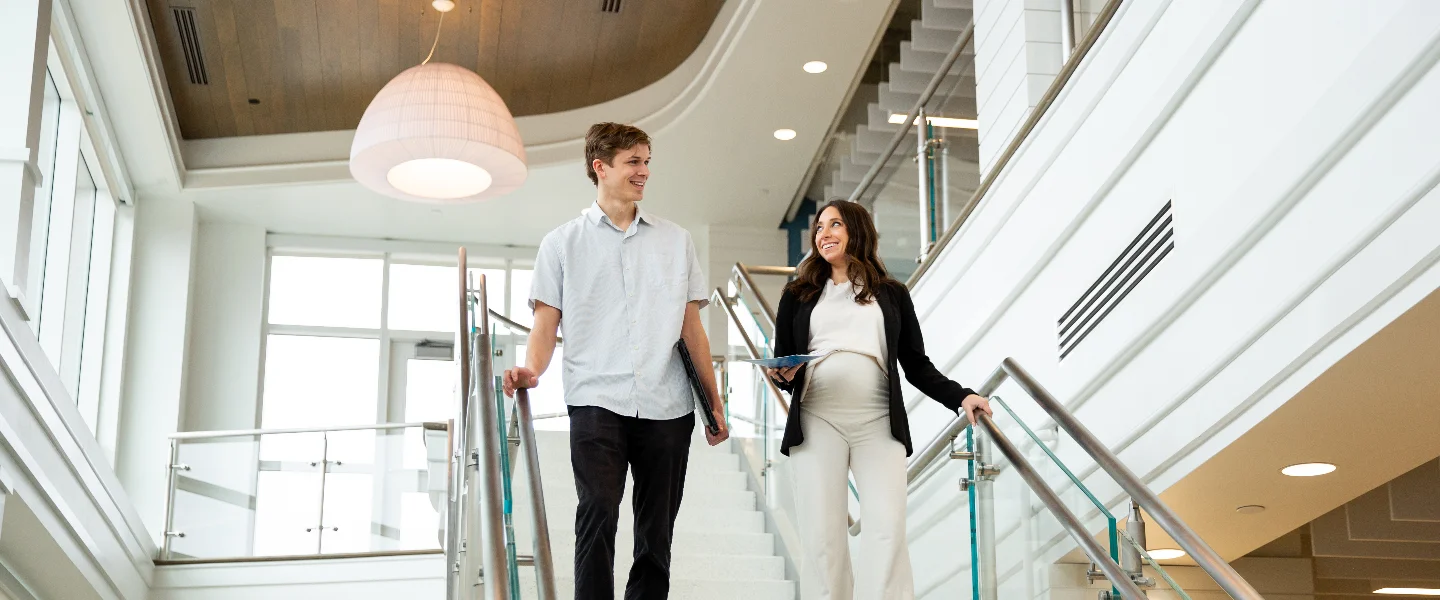 A man and a pregnant woman smile while walking down a modern office staircase, illustrating professional life.