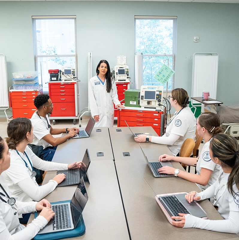Nursing students seated at a table listening to a nurse educator.