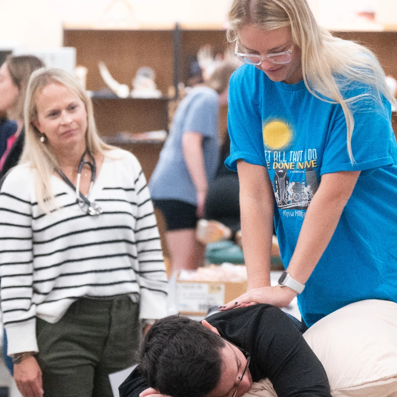 Student demonstrates a hands-on physical therapy technique on a classmate lying face down, while an instructor observes in a classroom setting.