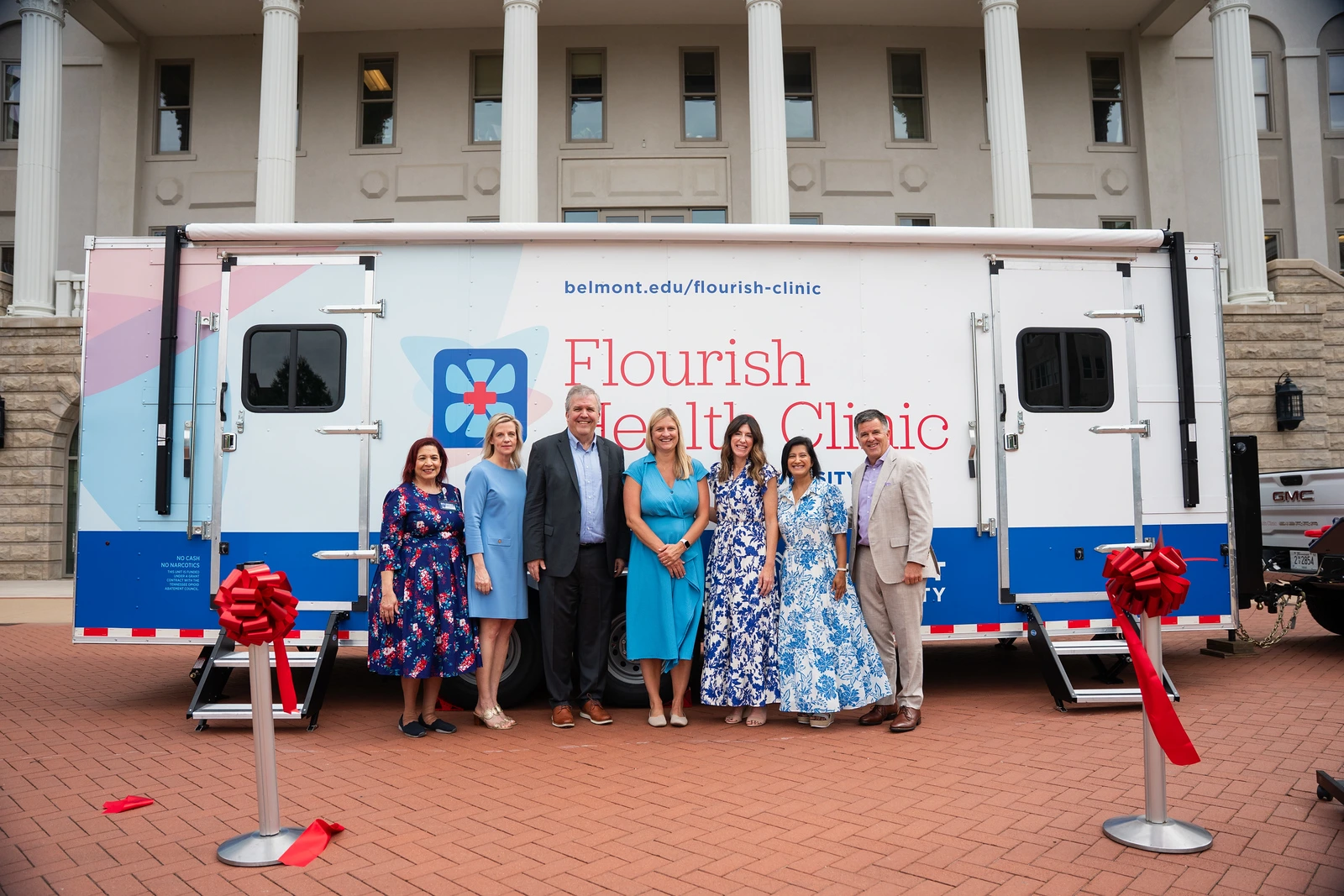 A group of seven people stands in front of a mobile clinic van labeled "Flourish Health Clinic" with red ribbon decorations, conveying a celebratory tone.