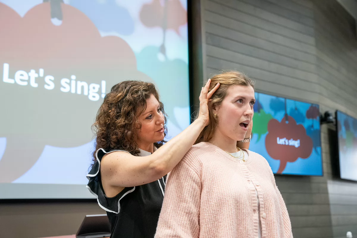 A Lindsay Ison instructs student in singing posture, gently touching her head. Background screens display "Let's sing!" in a bright, encouraging environment.
