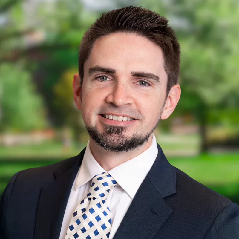 Portrait of a smiling professional man in a dark suit and patterned tie, standing outdoors against a softly blurred green park background.